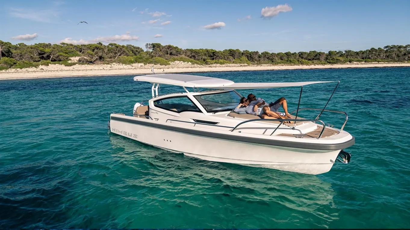 A couple relaxes on a Nimbus boat in clear turquoise waters, with sandy beach and trees in the background.
